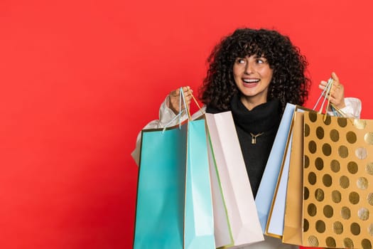 Happy Caucasian young woman with curly hair showing shopping bags, advertising discounts, smiling looking amazed with low prices, purchase on Black Friday holidays. Girl isolated on red background