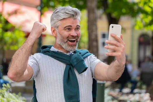 Happy Caucasian bearded man use mobile smartphone celebrating win good message news, lottery jackpot victory, online outdoors. Male middle-aged freelancer in city street. Business people lifestyle.