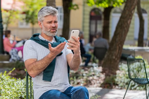 Bearded mature man using smartphone typing browsing, loses becoming surprised sudden lottery results, bad news, fortune loss, fail. Tourist guy standing in urban city street outdoors. Town lifestyles.