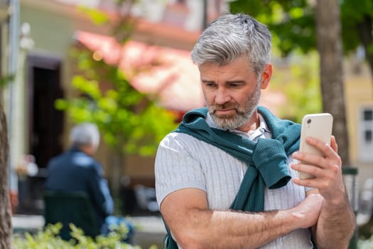 Bearded mature man using smartphone typing browsing, loses becoming surprised sudden lottery results, bad news, fortune loss, fail. Tourist guy standing in urban city street outdoors. Town lifestyles.