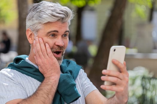 Oh my God, wow. Excited happy joyful winner mature man using smartphone looking at screen browsing celebrating win good message news. Caucasian guy traveler with mobile phone in urban street outdoors