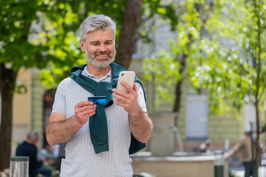 Happy mature Caucasian man using credit bank card smartphone while transferring money, purchases online shopping, order food delivery, booking hotel room. Guy standing in urban city street outdoors.