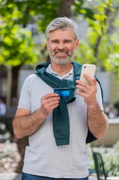 Happy mature Caucasian man using credit bank card smartphone while transferring money, purchases online shopping, order food delivery, booking hotel room. Guy standing in urban city street outdoors.