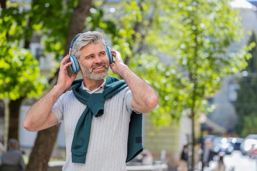 Happy mature Caucasian man in wireless headphones choosing, listening favorite energetic music in smartphone app outdoors. Bearded guy tourist walking passes by urban sunshine city street. Lifestyles