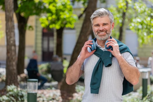 Joyful relaxed overjoyed mature bearded man listening favorite energetic disco rock n roll music dancing outdoors. Adult guy taking off wireless headphones looking at camera smiling on city street.