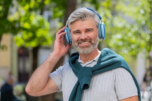 Happy mature Caucasian man in wireless headphones choosing, listening favorite energetic music in smartphone app outdoors. Bearded guy tourist walking passes by urban sunshine city street. Lifestyles