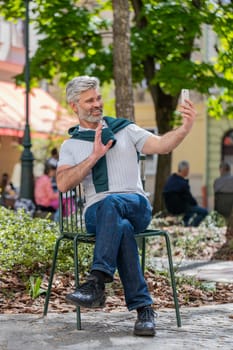 Bearded mature man blogger using smartphone, communicating video call online with subscribers, recording stories for social media vlog outdoors. Guy sitting on chair in urban sunshine city street.