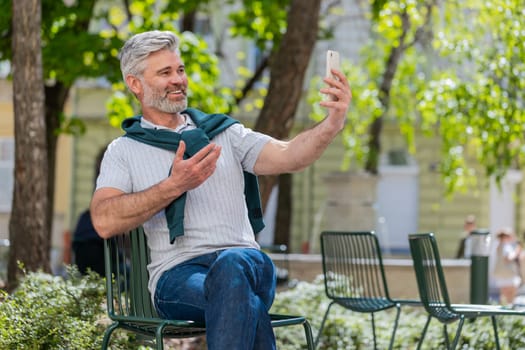 Bearded mature man blogger using smartphone, communicating video call online with subscribers, recording stories for social media vlog outdoors. Guy sitting on chair in urban sunshine city street.