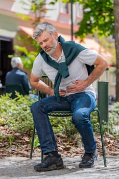 Caucasian mature man feeling sudden strong abdominal stomach ache, gastritis problem. Upset tourist guy having symptom poisoning diarrhea indigestion peptic ulcer pancreatitis on city urban street.