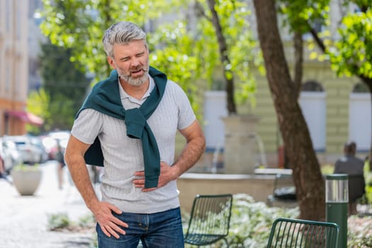 Caucasian mature man feeling sudden strong abdominal stomach ache, gastritis problem. Upset tourist guy having symptom poisoning diarrhea indigestion peptic ulcer pancreatitis on city urban street.