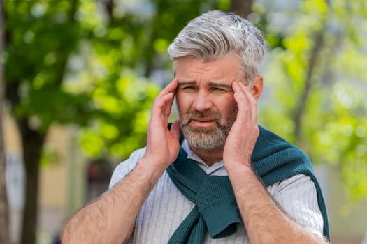 Displeased exhausted Caucasian man rubbing temples to cure headache problem suffering from tension migraine stress. Mature bearded guy tourist grimacing, pain standing on city urban street town.