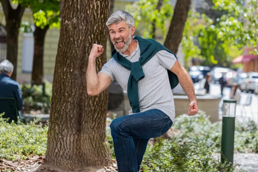 Excited happy man traveler shouting, celebrating success, winning, goal achievement, good news, lottery jackpot luck, victory outdoors. Mature guy walking on urban city sunshine street. Lifestyles.