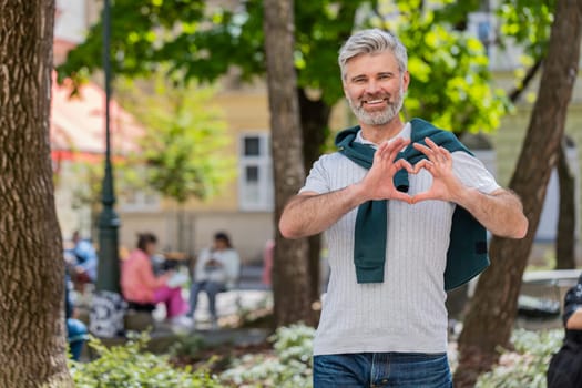 I love you. Caucasian middle-aged man makes symbol of love showing heart sign to camera, express sincere romantic positive feelings. Charity, gratitude, donation. Mature guy on urban city street park.