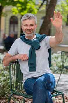 Happy mature man tourist smiling friendly at camera, waving hands gesturing invitation hello, hi, greeting, goodbye, welcoming with hospitable expression outdoors. Guy on urban city town street.