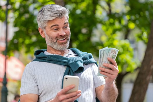 Happy rich mature man counting holding money dollar cash, use smartphone calculator app, plans to order gifts and food delivery online, booking hotel room. Bearded guy tourist in city street, outdoors