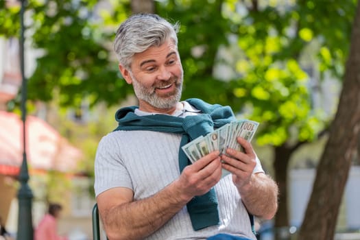 Rich happy mature man counting money cash, calculate domestic bills salary sitting on chair. Bearded guy satisfied of income earnings saves money for planned gifts. Bearded guy tourist on city street