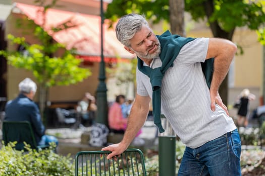 Caucasian mature man suffering backache, massaging sore place lower spine, frowning from unbearable spasm torn muscles and tendons, kidney stones pinched nerve. Bearded guy tourist in city street.