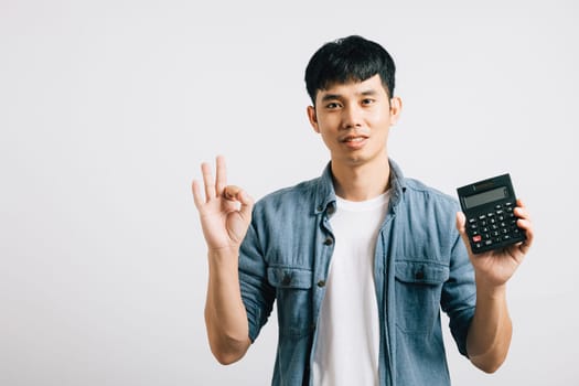 A TAX day concept comes to life with young man portrait on grey background. demonstrating a calculator, cash savings, and giving an OK sign with finger. symbolizes successful financial planning.