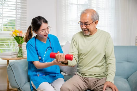 Young nurse physiotherapist helping old man in lifting dumbell at home, Asia nurse support elderly retirement sitting on sofa using red dumbbell workout exercise physical recovery, Health care concept