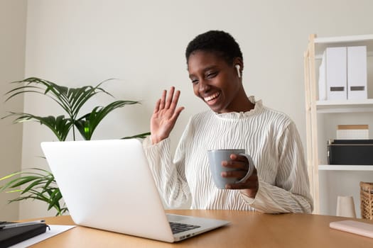Young Black woman waving hand on a video call at home office using wireless earphones and laptop. Working at home concept.