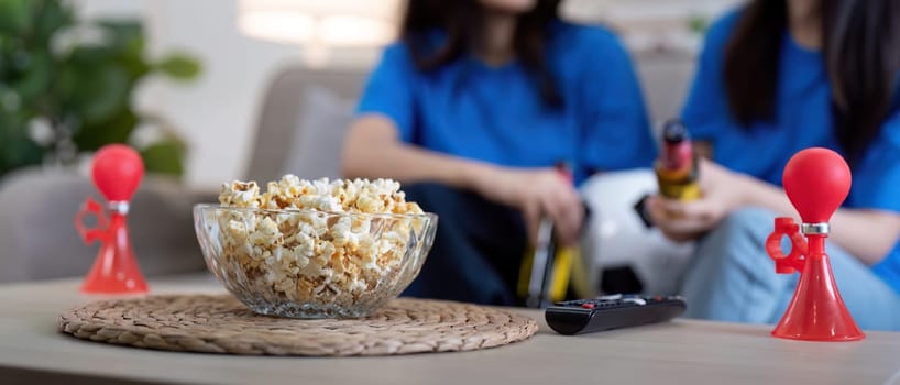 Lesbian couple cheering for Euro football at home with popcorn and air horn. Concept of LGBTQ pride, celebration, and sports excitement.