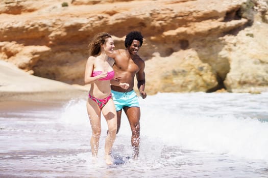 Full body of happy multiracial couple in swimwear walking in splashing water of sea and looking away while having fun together during sunny day