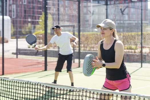 Portrait of positive young woman and adult man standing on padel tennis court, holding racket and ball, smiling. High quality photo