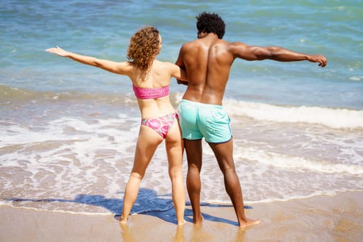 Full body back view of anonymous African American boyfriend and curly haired girlfriend in swimwear having fun near foamy sea during summer journey