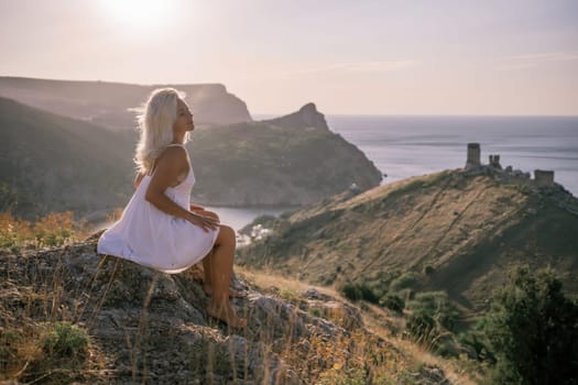 A woman is sitting on a hillside overlooking the ocean. She is wearing a white dress and has blonde hair. The scene is serene and peaceful, with the ocean in the background