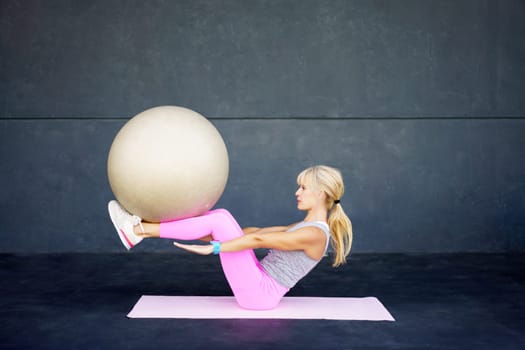 Full body side view of fit young female in sportswear exercising with fitness ball on yoga mat against gray wall in gym