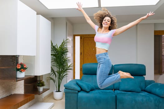 Happy young female wearing jeans and top jumping above ground near comfortable blue sofa in modern apartment with potted plants while looking at camera