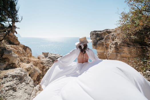 A woman in a white dress is standing on a rocky cliff overlooking the ocean. She is wearing a straw hat and she is enjoying the view