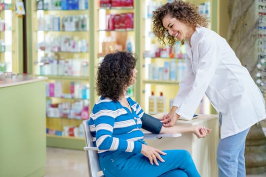 Smiling young female pharmacist in white robe, while standing near sitting lady customer with legs crossed and looking at each other during blood pressure checking near blurred shelves in pharmacy