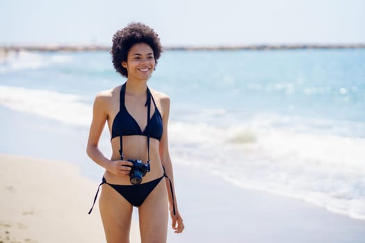 Optimistic African American female traveler in bikini with photo camera strolling on sandy beach near waving sea on sunny summer day