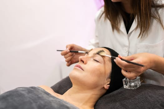 Crop cosmetician applying liquid cosmetic with brushes on face of female client lying in beauty salon during skin care treatment