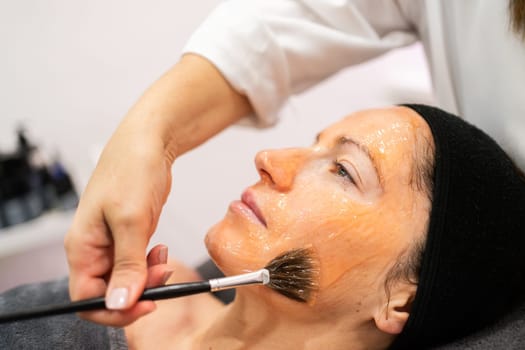 Crop professional beautician applying cosmetic gel on face of woman with brush while doing beauty treatment in modern salon during skin care routine