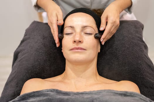 High angle of relaxed woman lying on couch under towel and getting professional skin care treatment in spa salon while cosmetician massaging face with crystals