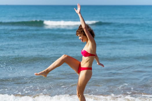 Side view of playful young female in swimwear walking in sea and kicking water while enjoying summer vacation at beach
