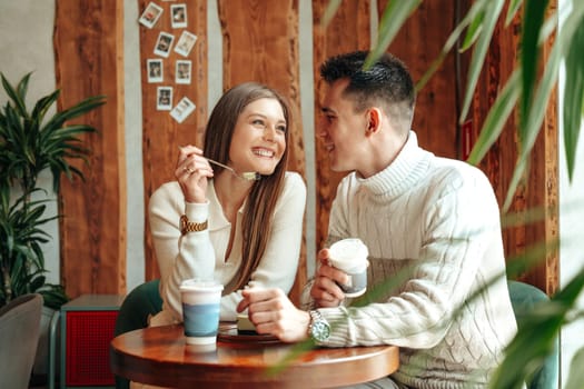 A young man and woman are seated at a small table in a warmly lit cafe, sharing a moment of laughter and connection. The woman, holding a coffee cup, playfully touches a fork to the mans nose, while he leans towards her with an amused expression. Surrounded by indoor plants and a casual, inviting ambiance, they appear to be enjoying a relaxed conversation over drinks.