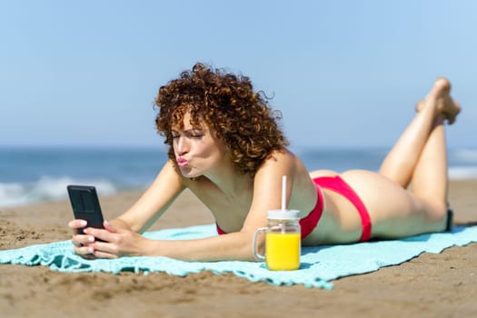Content female with curly hair in bikini pouting lips while taking selfie with smartphone and lying on beach with cool juice