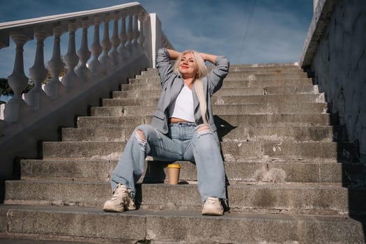 A woman in jeans and a jacket sits on a set of stairs. She is smiling and holding a cup