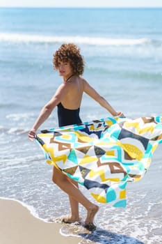 Full body side view of young curly haired female, with colorful flowing cloth behind standing on sandy seashore and looking behind back at camera in daylight