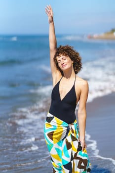 Smiling attractive young female in casual colorful clothes standing on wavy water seashore with raised arm and closed eyes on sunny day