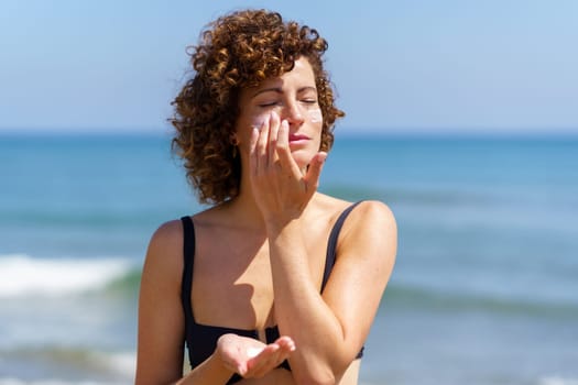 Sensual woman in bikini standing near waving ocean while applying sunblock cream on face during summer vacation