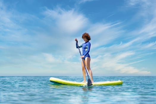 Full body of female in swimsuit paddling while standing on SUP board in sea against cloudy blue sky in sunlight