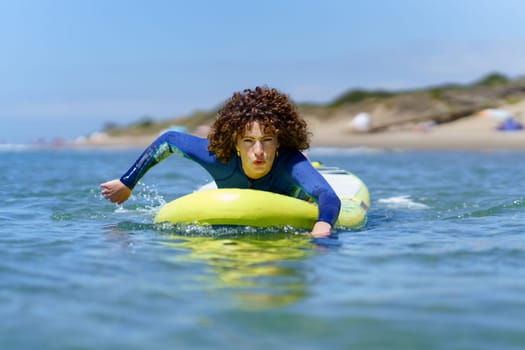 Concentrated young female surfer in wetsuit with curly hair lying on SUP board while paddling with hands floating on ocean during summer vacation
