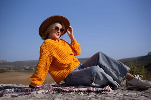 A woman in a yellow sweater and blue jeans is sitting on a blanket on a hillside. She is wearing sunglasses and a brown hat