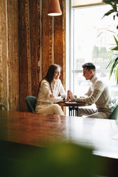 Two professionals are engaged in a conversation while sitting opposite each other at a wooden table in a cafe with natural light streaming in from a large window. They appear relaxed and focused, with cups of coffee in hand, surrounded by a warm, rustic interior.