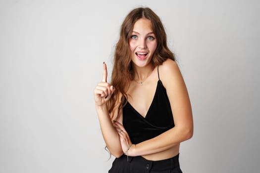 A smiling young woman with long wavy hair raises her index finger as if shes about to express an idea. She wears a black velvet dress and has a playful look on her face. The light grey background provides a neutral backdrop that highlights her figure.