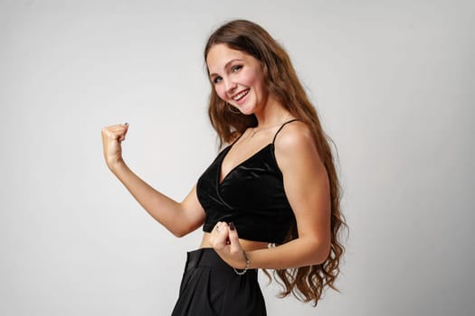 A young woman with long hair and a black top smiles brightly while flexing her arm muscle. She is standing against a neutral grey backdrop, exuding confidence and positive energy through her playful pose and cheerful expression.